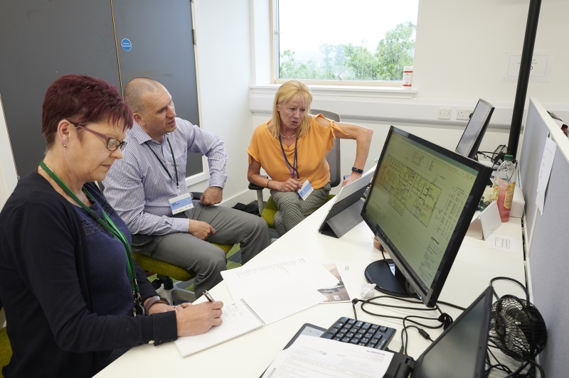 three people sitting a desk with laptop and computer screen 